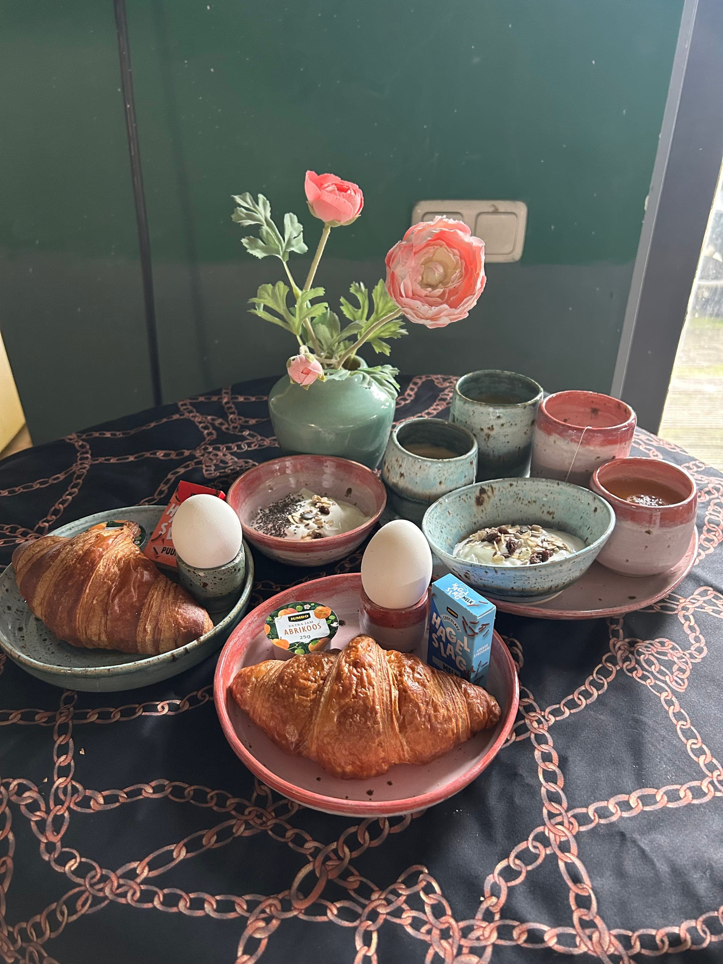 Ontbijt voor twee op geglazuurd servies: croissants, eitjes en yoghurt, met een vaasje ranonkels